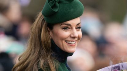 Kate Middleton wearing a green hat and coat smiling and holding flowers with a crowd of people blurred behind her