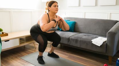 woman doing a squat on a grey yoga mat in a living room setting, with a grey sofa to the side of her. she&#039;s wearing black leggings, black trainers and a grey crop top.