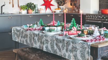 A dining table set up in the kitchen decorated for a Christmas dinner with a seasonal tablecloth, Christmas crackers, candlesticks and paper trees