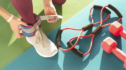 A person is tying an ankle weight around their ankle; their foot rests on a yoga mat. Nearby are resistance bands and a pair of dumbbells