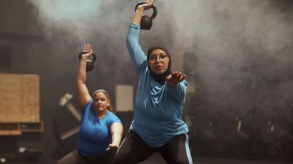 two women performing a kettlebell snatch facing the camera in a dark gym setting. 