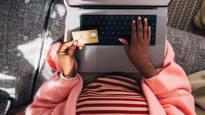 From above view of an Afro-American woman sitting on the sofa in her living room, shopping online using a credit card and laptop.