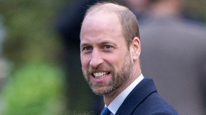 Prince William smiles while wearing a navy suit, he has a beard