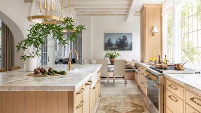 A briwn wooden kitchen with white marbel countertops, a large kitchen island, and gold fixtures. A large dining room at the end of the room