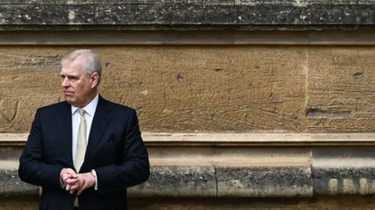 Prince Andrew wearing a dark suit and yellow tie standing in front of a tan concrete wall