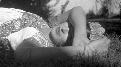 a woman (frida kahlo) wearing a flower crown lies in a grassy field while resting her arm over her eyes