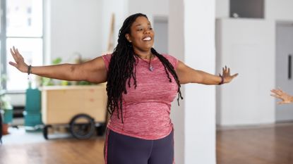 A woman in sportswear stands and stretches her arms out to either side. She is smiling; behind her we see white washed walls and wooden flooring.