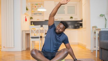 Man sitting cross-legged on the floor stretching one arm overhead and to the opposite side