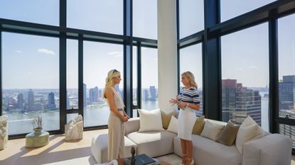 Taylor and Eleonora, standing in the living room of an NYC high-rise with floor-to-ceiling windows, in episode 108 of Selling the City.