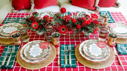 Christmas Table setting with a red and white tablecloth and matching napkins but in green and white. There is a woven charger placemat, and christmas plates stacked on top. The centerpiece is a red poppy arrangment.