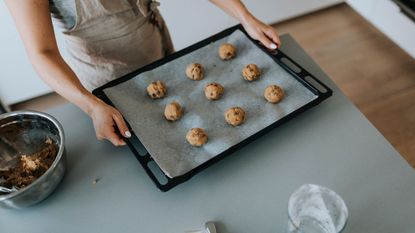 A Caucasian woman holding a black baking tray of cookies lined with baking paper