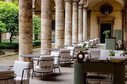 An ancient cloister hosts a chicly decorated cafe with round wrought iron tables and white upholstered outdoor furniture, as well as green-hued ceramic columns.