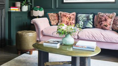 A dark green living room with a light pink velvet sofa, floral cushions and a coffee table displaying a flower vase and coffee table books