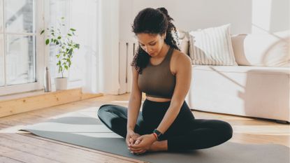 woman in a living room setting doing a seated butterfly hip stretch on a yoga mat. 