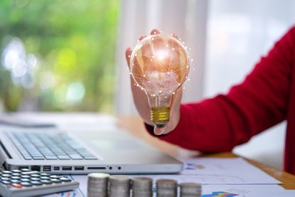 Hand holding a lightbulb above table with bills and money on it