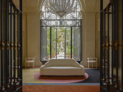 A palatial hotel&#039;s lobby features colorful carpet, a vintage-style white upholstered sofa, and a monumental Murano glass chandelier.