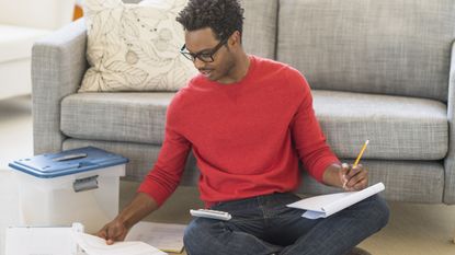 A smiling young man sits on the floor in front of his sofa and looks at paperwork with a calculator nearby.