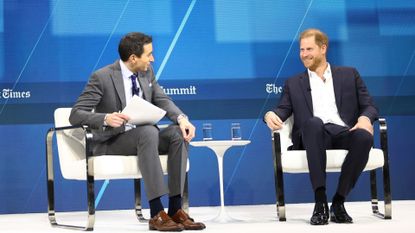 Prince Harry wearing a blue suit and sitting on a white carpeted stage with Andrew Ross Sorkin