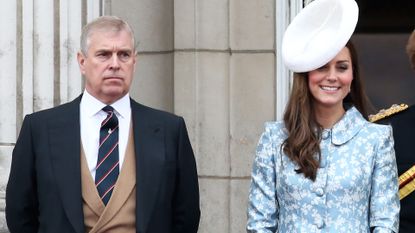 Prince Andrew wears a suit, waistcoat, and tie while stranding on the Buckingham Palace balcony next to Kate Middleton who wears a turquoise blue and white floral outfit and a large white hat