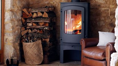 Close up a log burner in a stone fireplace beside a leather armchair with a log pile to show the best wood for log burners