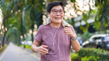 woman wearing red rimmed glasses and lilac tshirt walking dynamically on a paved path through trees. She has a small towel around her neck, earphones in and a wearing a fitness watch