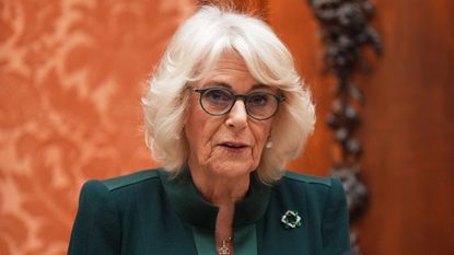 Queen Camilla wearing a green dress and brooch with glasses standing in front of an orange wall