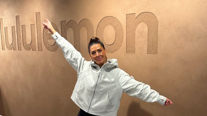 Woman in white jacket stands in front of brown wall with lululemon embossed on it