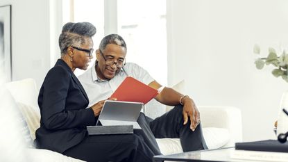 An older couple look at a folder of paperwork together on the sofa.