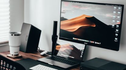 MacBook Pro sitting on a desk beneath a black external monitor, with a coffee cup to the side