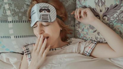 A woman with a sleep mask on her head while yawning in bed