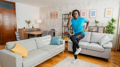 A man marches on the spot in a living room, wearing a t-shirt, joggers and socks. He swings his arms as he walks on the spot, lifting his knees up high. Surrounding him in the room, we see two couches, a table and chairs, decorative shelving, leafy plants and several pictures hung on the walls.