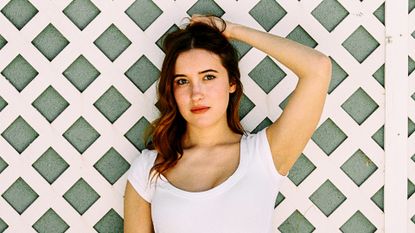 sex lives of college girls star gracie lawrence poses with her arm above her head in a white tshirt by a fence