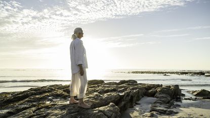 An older woman stands on some rocks at the beach and looks into the distance.