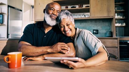 An older couple look happy and confident while looking at a tablet at their kitchen table.