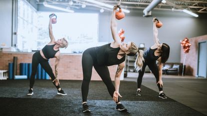 Three women performing the kettlebell windmill exercise in a gym. They bend to the side at the waist and hold a kettlebell above them.