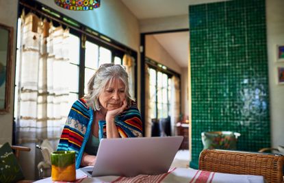 Senior woman wrapped in blanket standing at dining table looking at emails on laptop