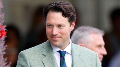 King Charles&#039; equerry Jonny Thompson smiles while wearing a light green suit, a white shirt, and a striped tie