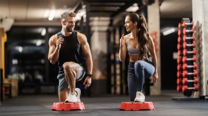 A man and woman perform a reverse lunge on a stepper in a gym setting. They are wearing sportswear and looking at each other. Their front knee is bent while the other is behind them. In the background we see a rack of dumbbells.