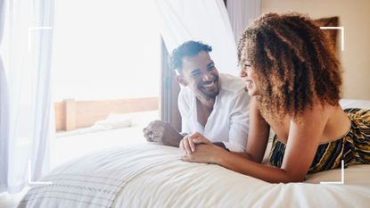 Man and woman lying on bed together fully clothed laughing and smiling, looking at each other, to represent the prone bone sex position