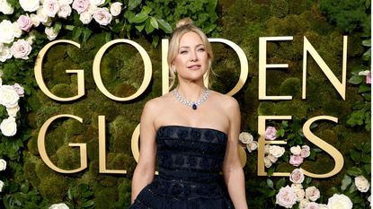 Kate Hudson in a black strapless dress standing in front of a green, moss and floral background that has the Golden Globes logo in large gold letters on it