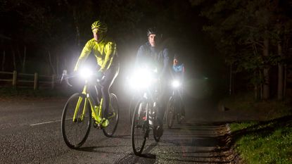 Four cyclists riding through the night, with lights at full beam. One is dressed in dark clothes and one in full fluorescent jacket, helmet and bike. In a 2 x 2 format. 