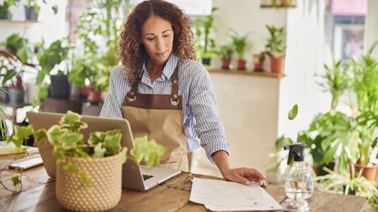 Small business owner of a nursery looks over paperwork while working on her laptop.