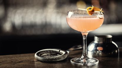 side shot of a coupe filled with a light orange cosmopolitan with a strainer on the countertop next to the drink