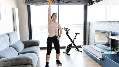 A woman exercises with a dumbbell at home. She is standing and holding the weight overhead, with a straightened arm; her other hand rests on her hip. Behind her is an exercise bike and large window. To either side of her is a couch and mounted television.