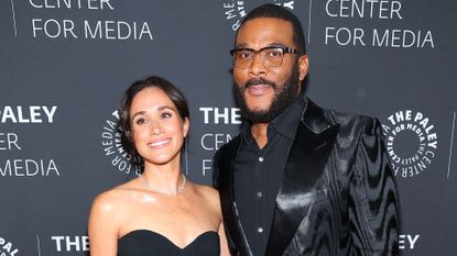 Meghan Markle wears a black strapless dress while posing with Tyler Perry who is dressed in a black suit and shirt