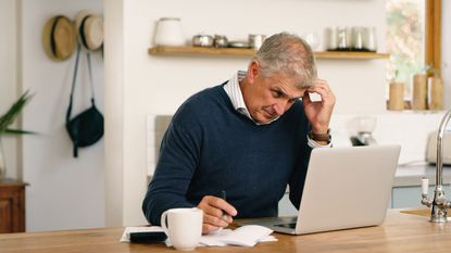 An older man looks a little frustrated as he looks at his laptop on his kitchen island while working on paperwork. 