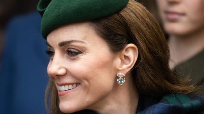 Kate Middleton wearing a blue and green plaid scarf and green hat looking to the left and smiling