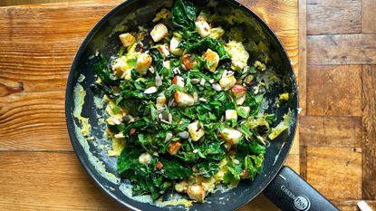 Black frying pan filled with green veg, scrambled eggs and cubes of halloumi on wooden table