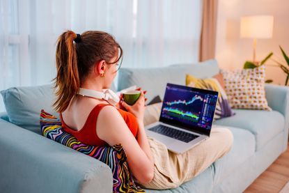 Young woman sitting on the sofa, looking at a laptop screen showing share price data