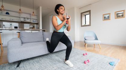 Woman performing Bulgarian split squat at home, with her rear foot elevated on a sofa, and one end of dumbbell held in front of her chest in both hands.